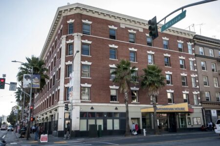 A four-story red-brick building with white trim stands at a street corner, with palm trees lining the sidewalk and people walking by. The ground floor has businesses, including one with a yellow “SAVANNAH HOUSE” sign.