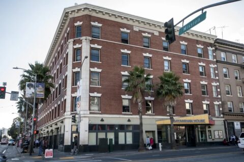 A four-story red-brick building with white trim stands at a street corner, with palm trees lining the sidewalk and people walking by. The ground floor has businesses, including one with a yellow “SAVANNAH HOUSE” sign.