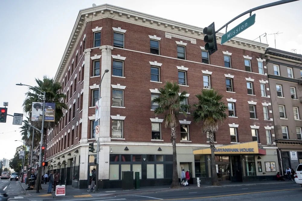 A four-story red-brick building with white trim stands at a street corner, with palm trees lining the sidewalk and people walking by. The ground floor has businesses, including one with a yellow “SAVANNAH HOUSE” sign.