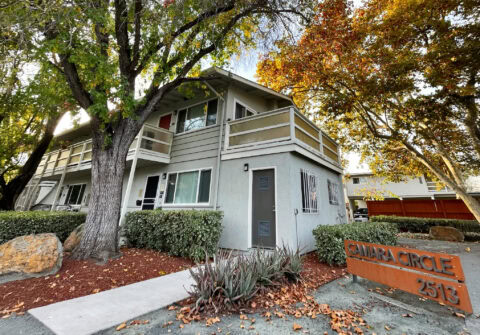 A two-story apartment building with balconies, surrounded by trees with autumn leaves. There is a sidewalk, some shrubs, and a sign in front reading "CAMARA CIRCLE 2513.