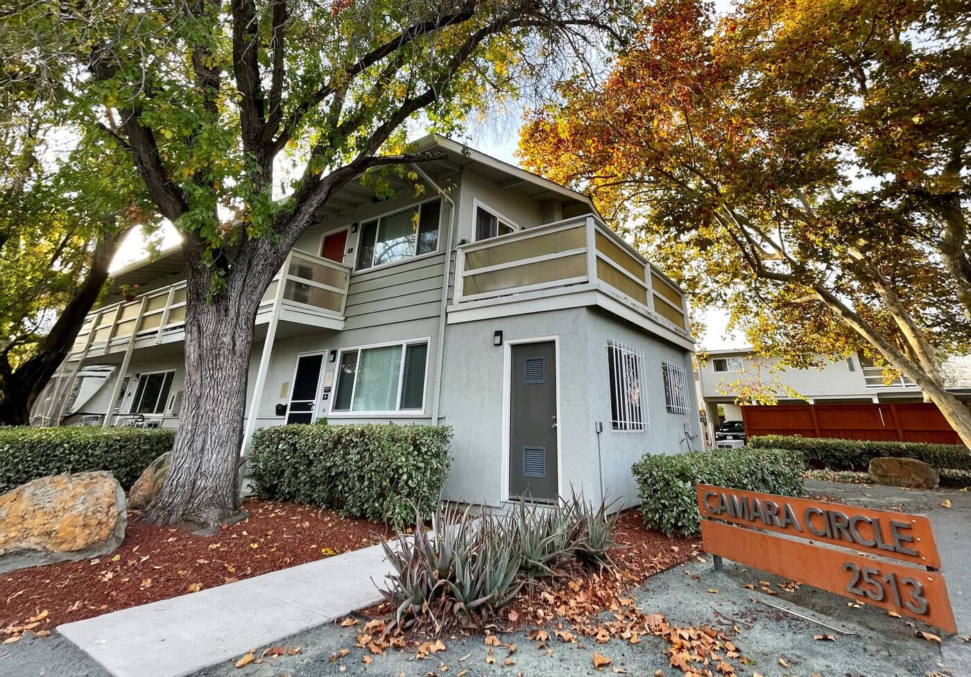 A two-story apartment building with balconies, surrounded by trees with autumn leaves. There is a sidewalk, some shrubs, and a sign in front reading "CAMARA CIRCLE 2513.