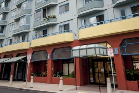 A modern apartment building with blue-trimmed windows, yellow balconies, and a red lower facade. There are glass awnings above the entrances and street signs along the sidewalk.