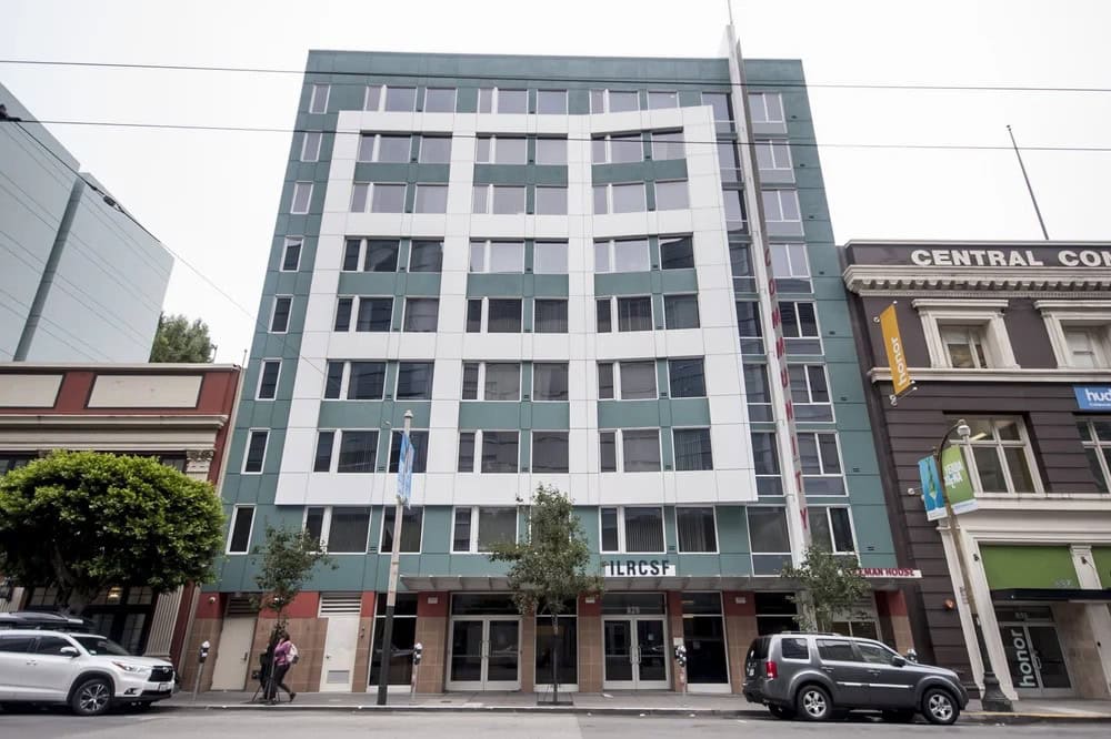 A modern, multi-story apartment building with a geometric white and green facade, large windows, and trees along the sidewalk. Cars are parked on the street in front. Adjacent older buildings are visible on both sides.