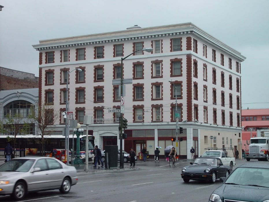 A four-story building with white and reddish-brown trim stands on a busy city corner. Cars drive by, people walk on sidewalks, and the sky is overcast, giving the scene a gray tone.