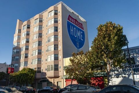 A multi-story beige apartment building with a large mural reading "INNER CITY HOME" on its side. Trees line the street, and several parked cars are visible in front of the building.
