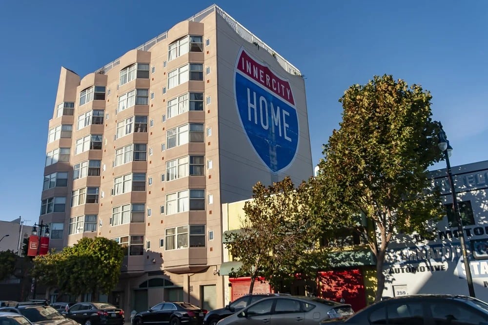 A multi-story beige apartment building with a large mural reading "INNER CITY HOME" on its side. Trees line the street, and several parked cars are visible in front of the building.