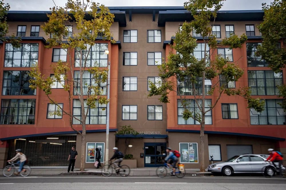 An apartment building with orange and brown exterior, large windows, and trees in front. Several people ride bicycles along the street, and a parked car is visible on the right side.
