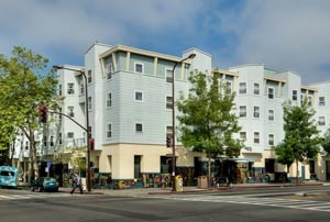 A four-story modern apartment building with white siding and large windows sits on a street corner, surrounded by trees, parked cars, and a few small businesses at street level.