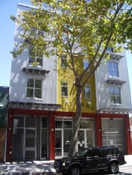 A modern four-story building with a yellow vertical accent, large windows, and a tree in front. A black SUV is parked on the street below under a sunny sky.