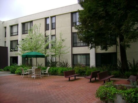 A courtyard with red brick flooring features benches, a table with chairs, and a green umbrella. There are trees, plants, and a three-story beige building with many windows in the background.