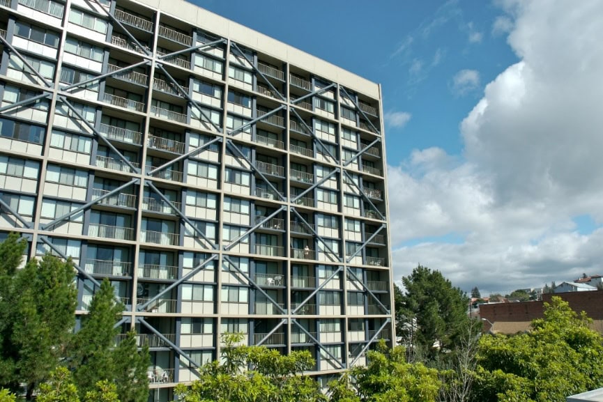 A modern apartment building with crisscrossing metal beams on its facade stands next to green trees, under a partly cloudy blue sky.
