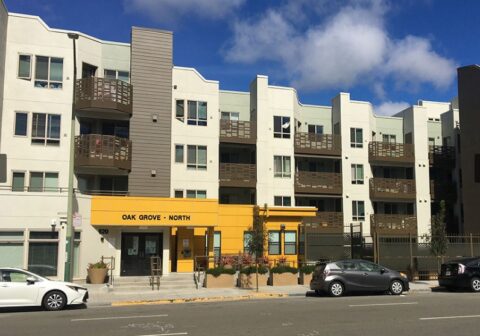 A modern four-story apartment building with balconies and a yellow entrance labeled "Oak Grove - North." Several cars are parked along the street in front of the building under a blue sky with clouds.