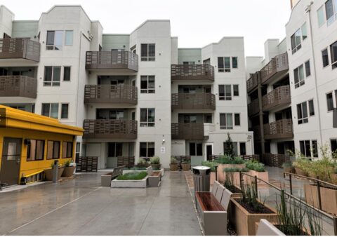 A modern apartment complex with multiple balconies, beige and brown exterior, and a central courtyard featuring benches, planters, and a yellow building to the left, all on a wet pavement.