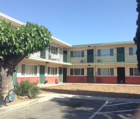 A two-story motel with green doors and railings, red brick lower walls, and a large leafy tree in the foreground under a clear blue sky.