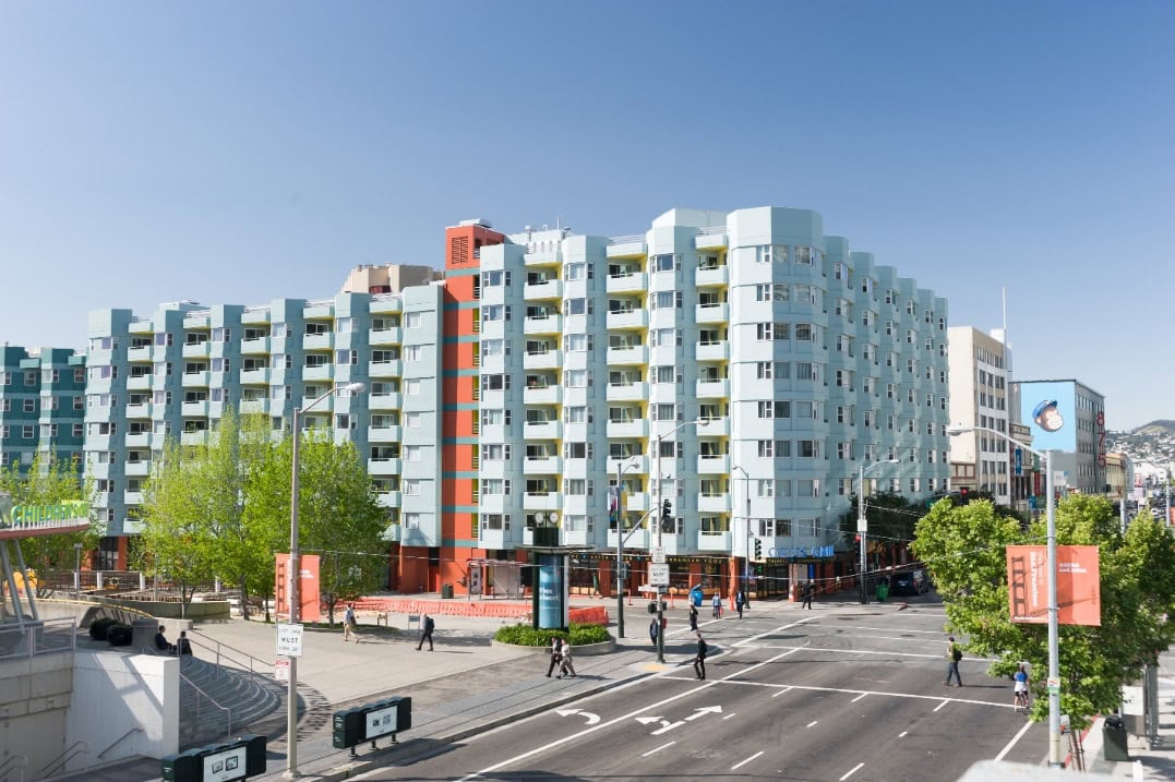 A large, modern light blue apartment building with orange accents stands at a busy city intersection, with people walking on the sidewalks and trees and clear blue sky in the background.