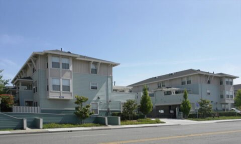Two modern, light blue apartment buildings with white trim stand along a quiet street under a clear blue sky. Small trees and shrubs line the sidewalk in front of the buildings.