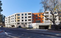 A modern, multi-story apartment building with white and orange sections, large windows, and an underground parking entrance, surrounded by trees on a clear, sunny day.