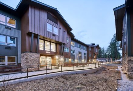 Modern multi-story apartment building with large windows, wood and stone accents, and a ramp leading to the entrance; pine trees and other buildings are visible in the background at dusk.