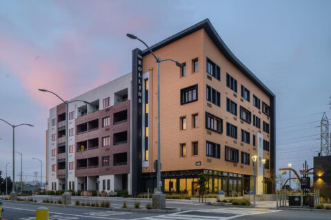 Modern five-story apartment building with tan and white exterior, large windows, balconies, and a sign reading "ROLLINS" on the corner, set at an intersection during dusk with streetlights and a pinkish sky.