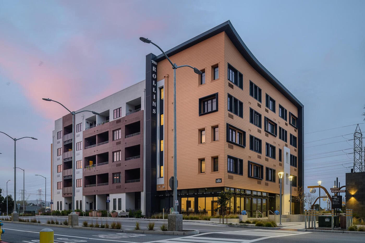 Modern five-story apartment building with tan and white exterior, large windows, balconies, and a sign reading "ROLLINS" on the corner, set at an intersection during dusk with streetlights and a pinkish sky.