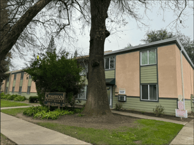 A two-story apartment building with beige and green exterior, large windows, and a tree in front. A sign reads “Crosswood Apartments, 4641 3rd Street E.” Shrubs and grass line the sidewalk.