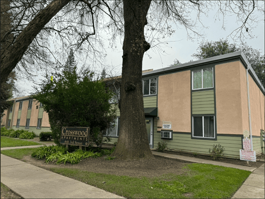 A two-story apartment building with beige and green exterior, large windows, and a tree in front. A sign reads “Crosswood Apartments, 4641 3rd Street E.” Shrubs and grass line the sidewalk.