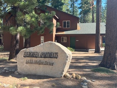 A large stone with "Evergreen Apartments, 1432 Melba Drive" engraved on it sits in front of a brown, two-story building surrounded by tall pine trees.