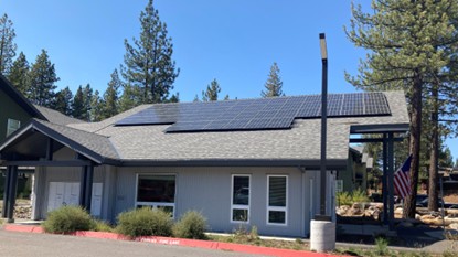 A small building with gray siding and a sloped roof has solar panels installed on top. There are large windows, a flagpole with an American flag, and trees in the background under a clear sky.