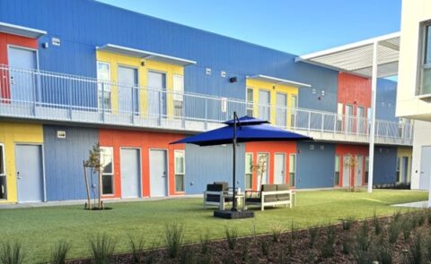 A modern apartment building with blue, yellow, and red accents surrounds a courtyard featuring artificial grass, a seating area, and a blue umbrella, with small trees and plants along the edge.