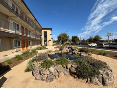 A low-rise apartment building with balconies overlooks a small landscaped pond surrounded by rocks and greenery under a sunny, blue sky with some clouds. Cars are parked along the adjacent street.
