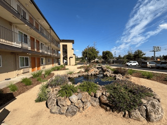 A low-rise apartment building with balconies overlooks a small landscaped pond surrounded by rocks and greenery under a sunny, blue sky with some clouds. Cars are parked along the adjacent street.