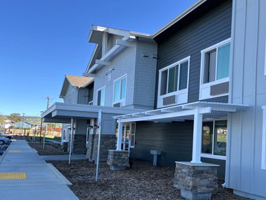 A modern two-story building with gray siding, white trim, large windows, and covered entrances supported by stone columns, under a clear blue sky. A sidewalk and mulch landscaping run along the front.