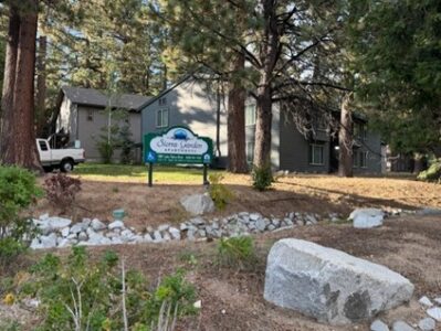 A sign for Sierra Garden Apartments stands among trees and rocks, with two gray apartment buildings and a white truck visible in the background. Pine needles and greenery cover the ground.