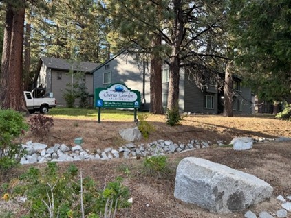 A sign for Sierra Garden Apartments stands among trees and rocks, with two gray apartment buildings and a white truck visible in the background. Pine needles and greenery cover the ground.