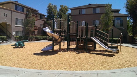 A playground with a slide, climbing structures, and a green dinosaur-shaped play feature on wood chip ground, surrounded by apartment buildings and trees on a sunny day.