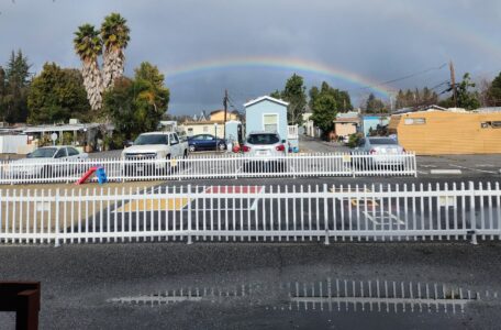 A rainbow arches over a parking lot with several cars, white picket fences, and a few houses and trees under a partly cloudy sky. Wet ground and small puddles reflect the scene.