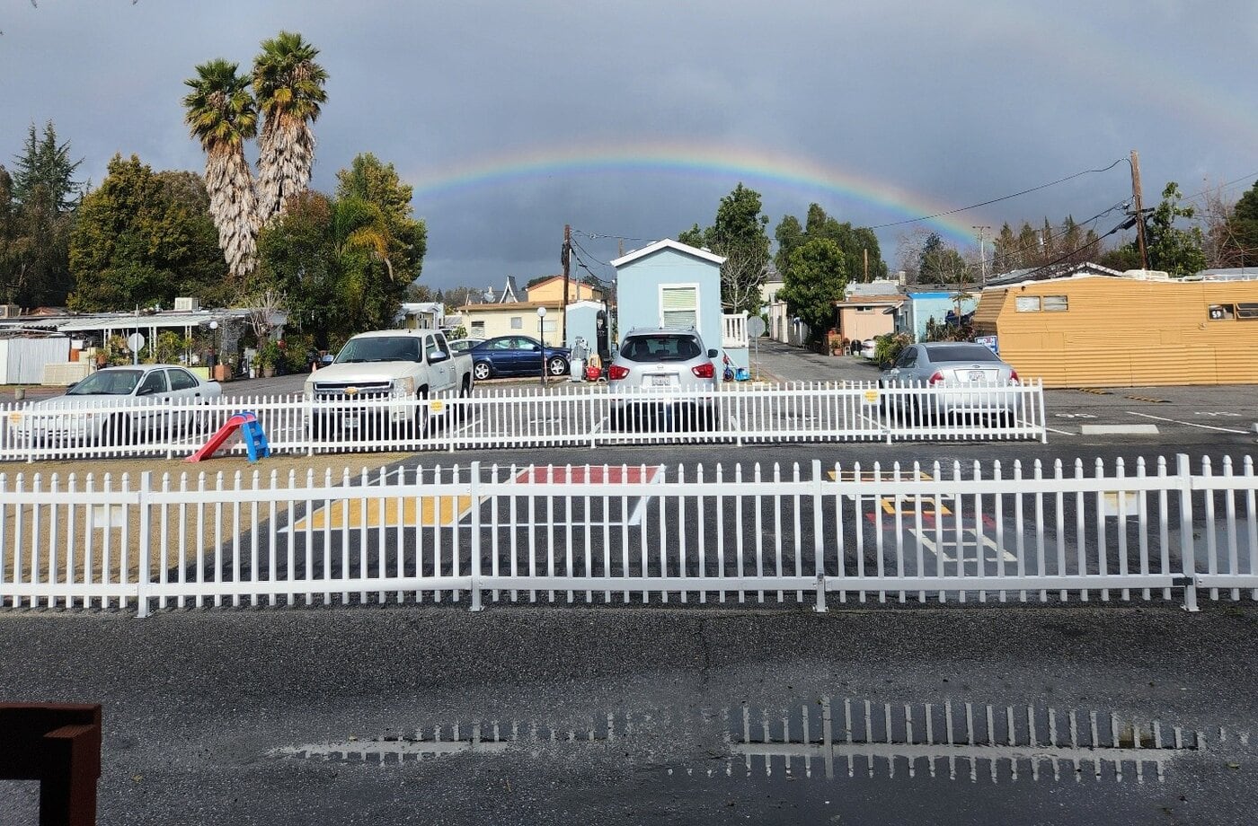 A rainbow arches over a parking lot with several cars, white picket fences, and a few houses and trees under a partly cloudy sky. Wet ground and small puddles reflect the scene.