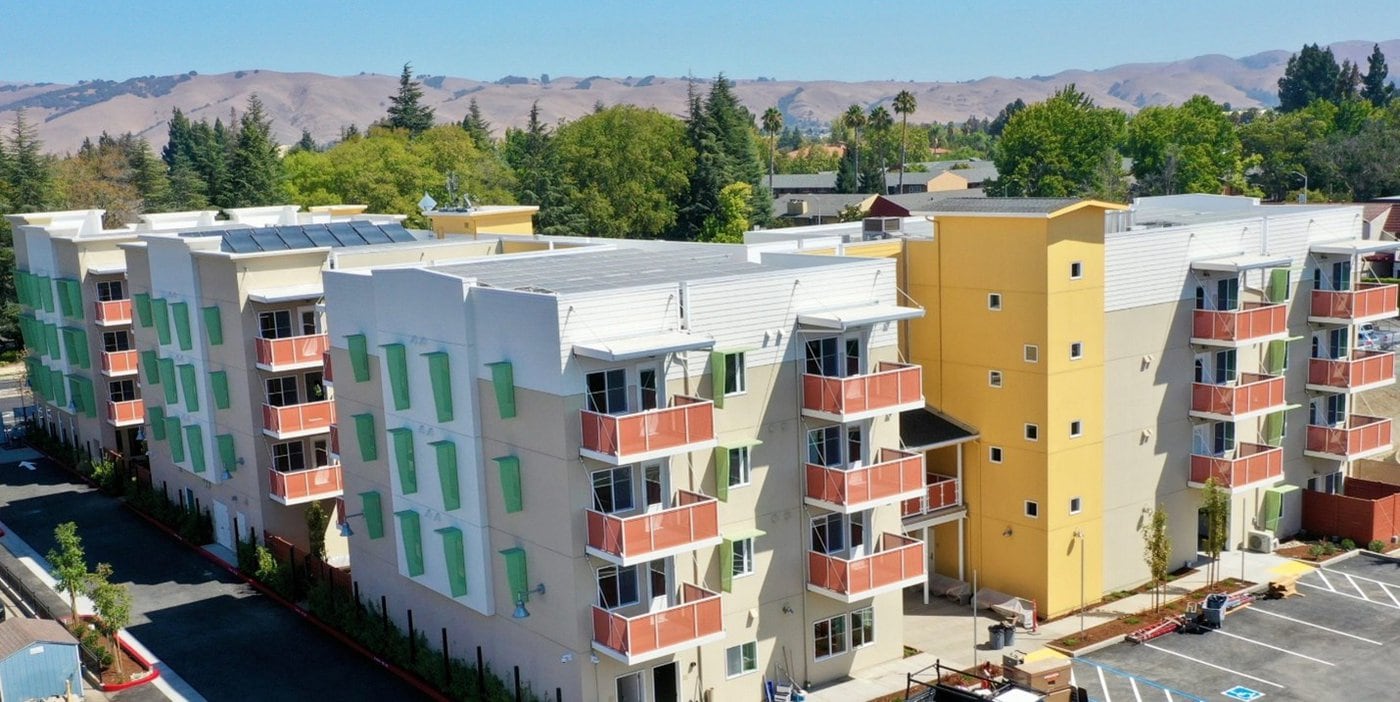 A modern, multi-story apartment building with colorful green and yellow accents, surrounded by trees and parked cars, with hills visible in the background under a clear sky.