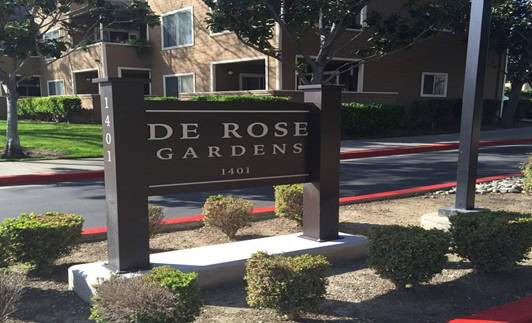 A dark brown sign with white letters reads "DE ROSE GARDENS 1401" in front of an apartment building, surrounded by trimmed bushes, trees, and a red curb.