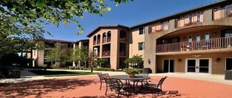 A sunny courtyard with metal patio tables and chairs in front of a modern three-story apartment building with balconies, large windows, and trees along the walkway.