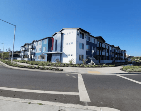 A modern, two-story apartment building with blue and white siding sits on a street corner under a clear blue sky. The landscaping includes green shrubs and small trees along the sidewalk.