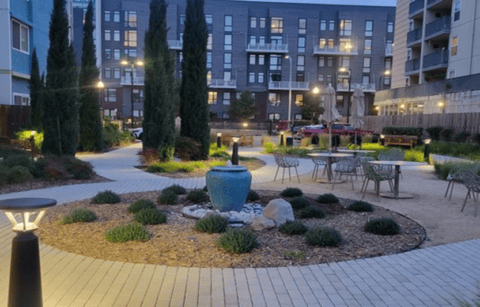 A landscaped courtyard at dusk features a large blue ceramic fountain in the center, surrounded by small plants, outdoor seating, trees, and apartment buildings in the background, softly lit by pathway lights.