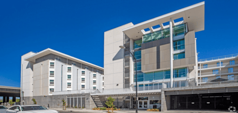 A modern multi-story apartment building with large windows, white walls, and a covered entrance. There is a car parked in front and a clear blue sky overhead.