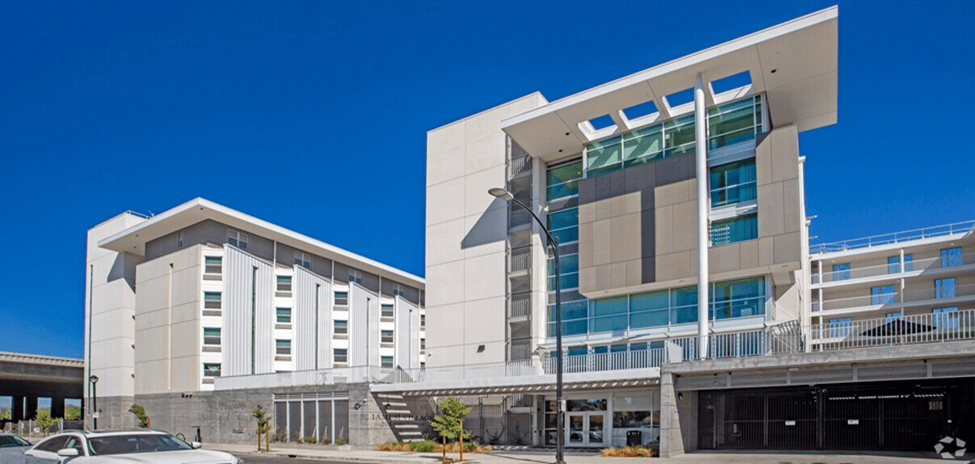 A modern multi-story apartment building with large windows, white walls, and a covered entrance. There is a car parked in front and a clear blue sky overhead.