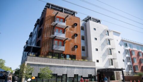 A modern multi-story apartment building with a mix of rust-colored and white facades, metal balconies, large windows, and solar panels on the rooftop, under a clear blue sky.