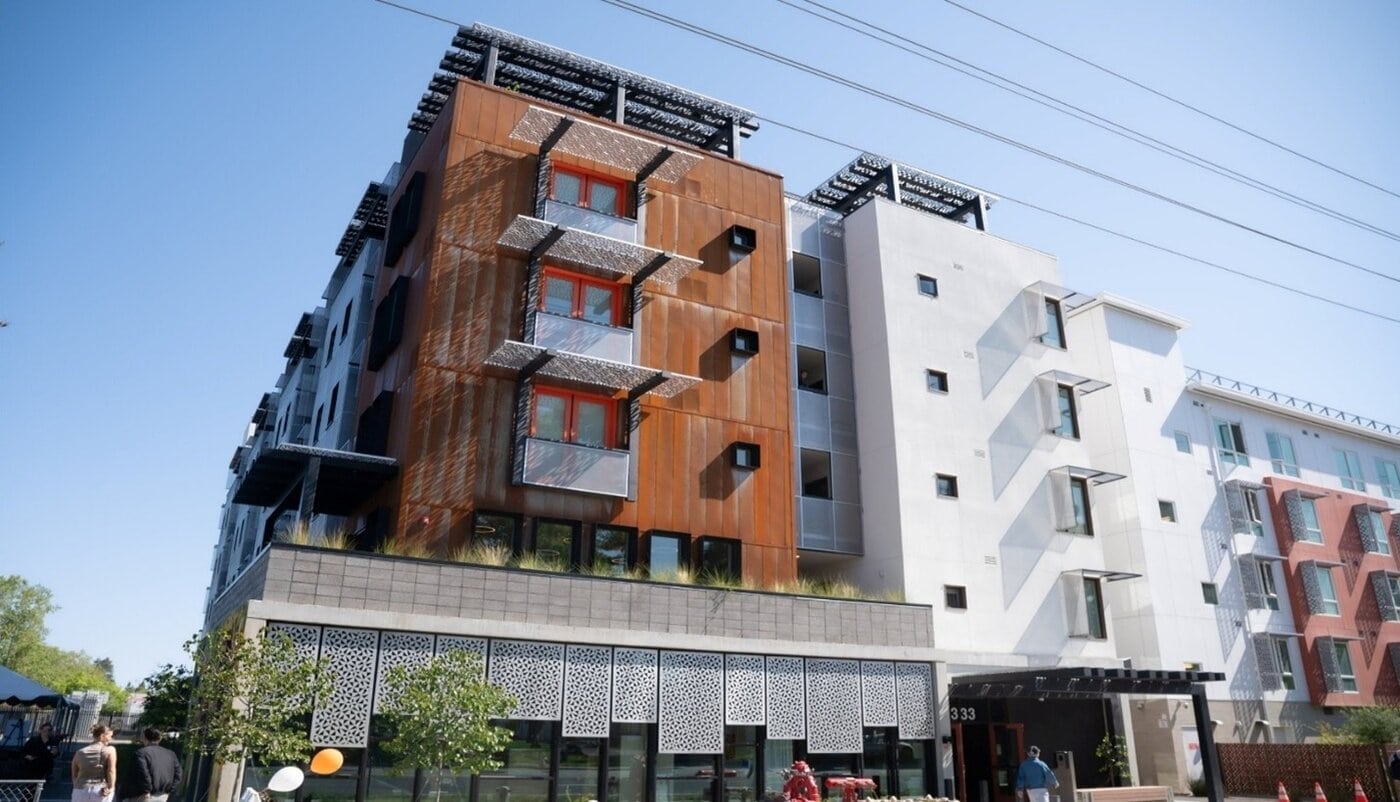 A modern multi-story apartment building with a mix of rust-colored and white facades, metal balconies, large windows, and solar panels on the rooftop, under a clear blue sky.