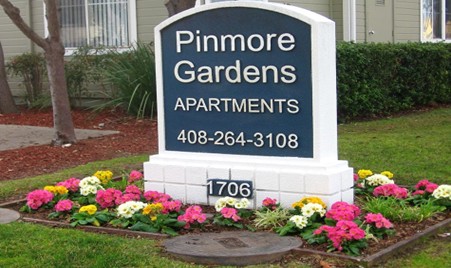 A sign for Pinmore Gardens Apartments with a phone number, surrounded by green grass and colorful flowers, located at 1706.