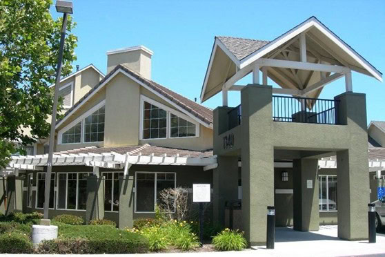 A modern building with large windows, gabled roofs, and a covered entryway with white trim, surrounded by green landscaping and a clear blue sky.