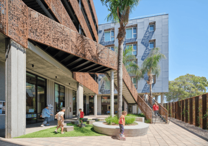 Children play and adults walk near a modern building with perforated metal panels, a staircase, palm trees, and large windows on a sunny day.