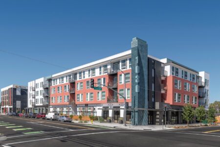 A modern four-story apartment building with red and white exterior, large windows, balconies, and a tall blue-gray vertical sign displaying 